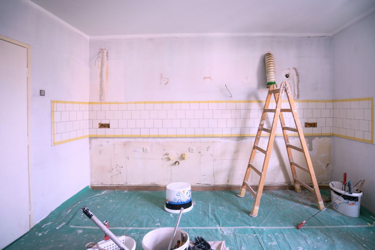 Kitchen undergoing renovation with cabinets removed, a ladder, paint buckets, and protected flooring.